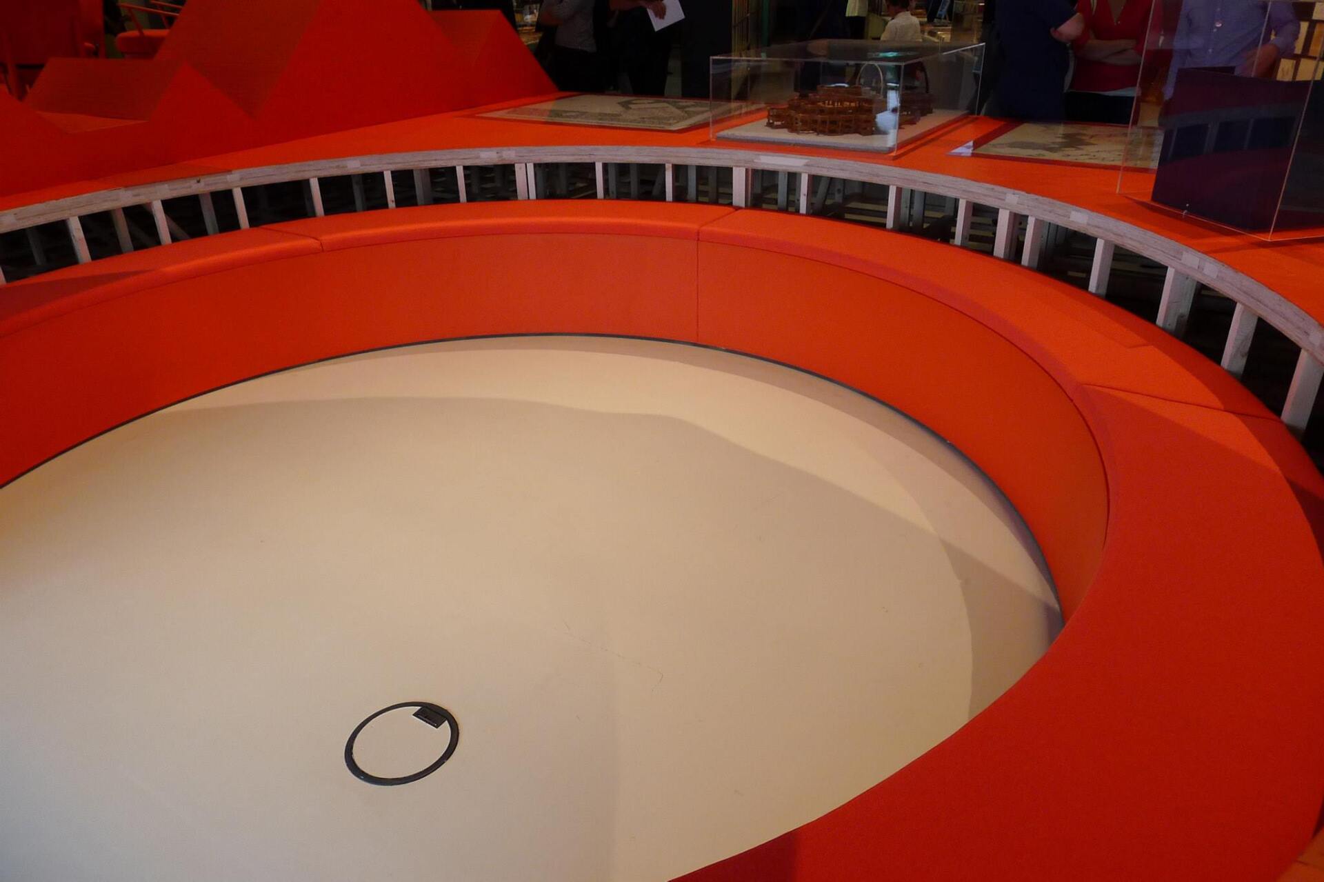 Ronde oranje structuur met een wit interieur, mogelijk een tentoonstellingsobject, met een grijs cirkelontwerp.