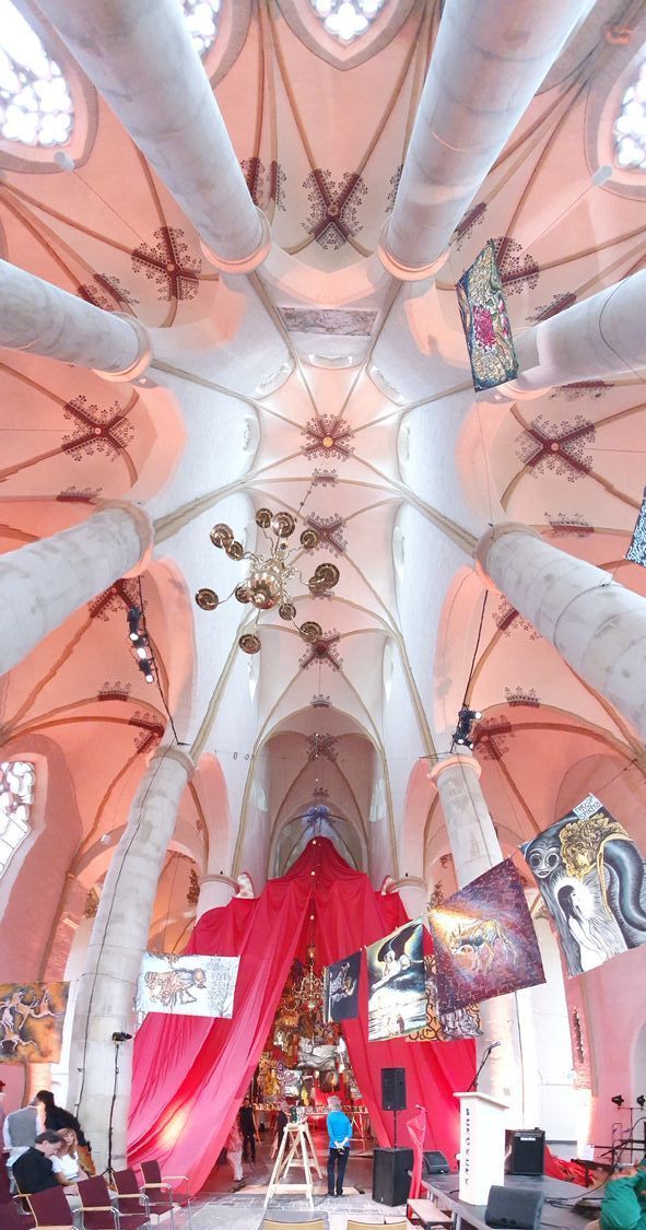 Binnen in een kerk, omhoog kijkend. Versierd plafond met pilaren en versieringen; rode draperieën onderaan, mensen zichtbaar.