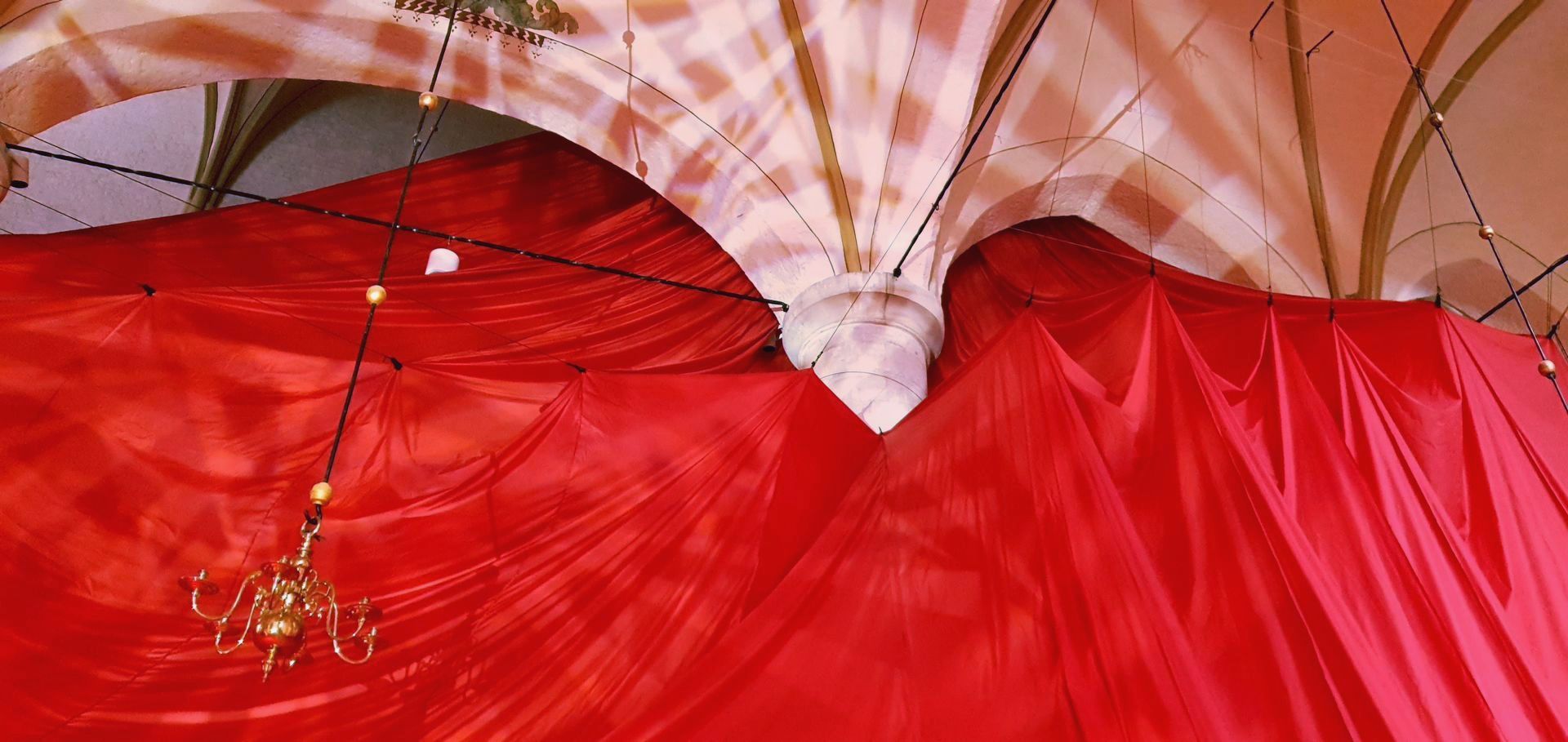 In een kamer met gewelfd plafond hangt een rode doek, waarin een decoratieve kroonluchter zichtbaar is.