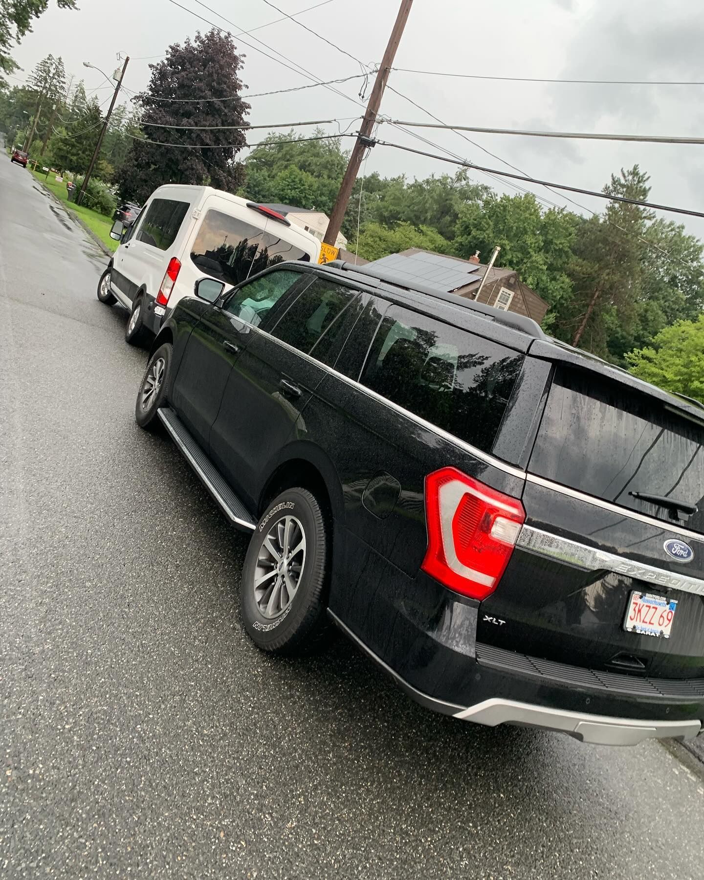 Black SUV and white van parked on a wet, sloped street next to a utility pole.