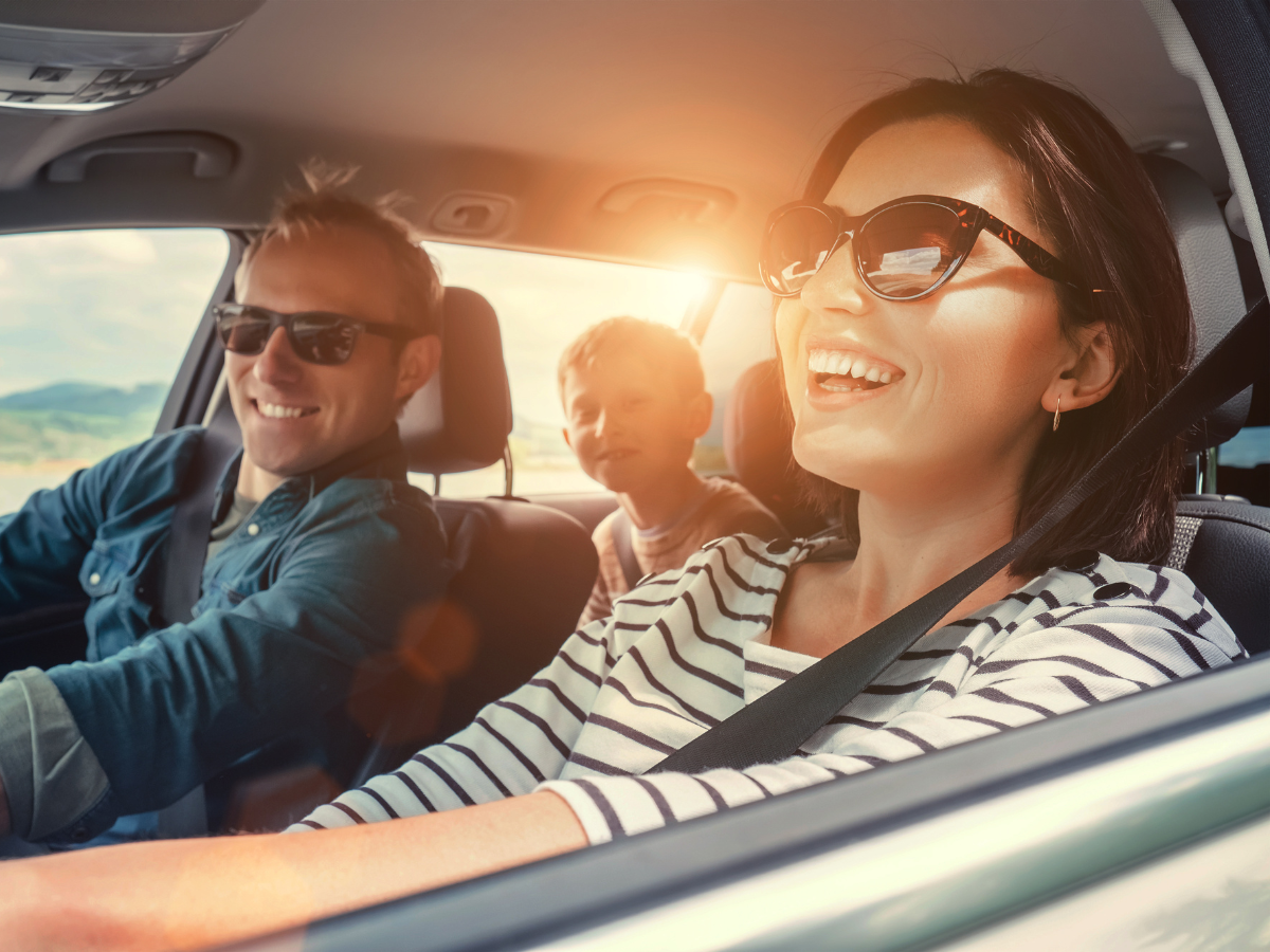 Family smiling in a car, sunlit interior. Driving on a road with mountains in the background.