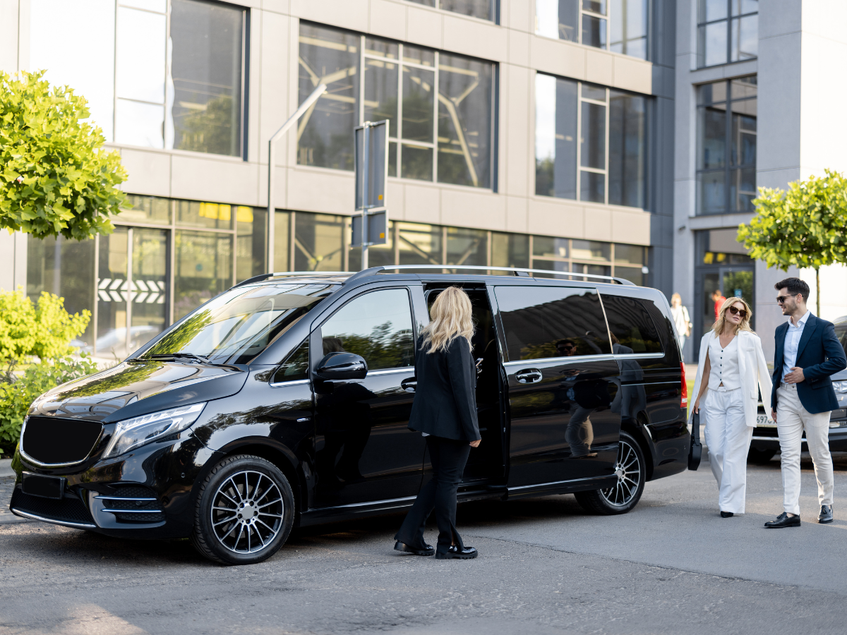Black van parked outside building; woman opens door for couple dressed in white and blue.