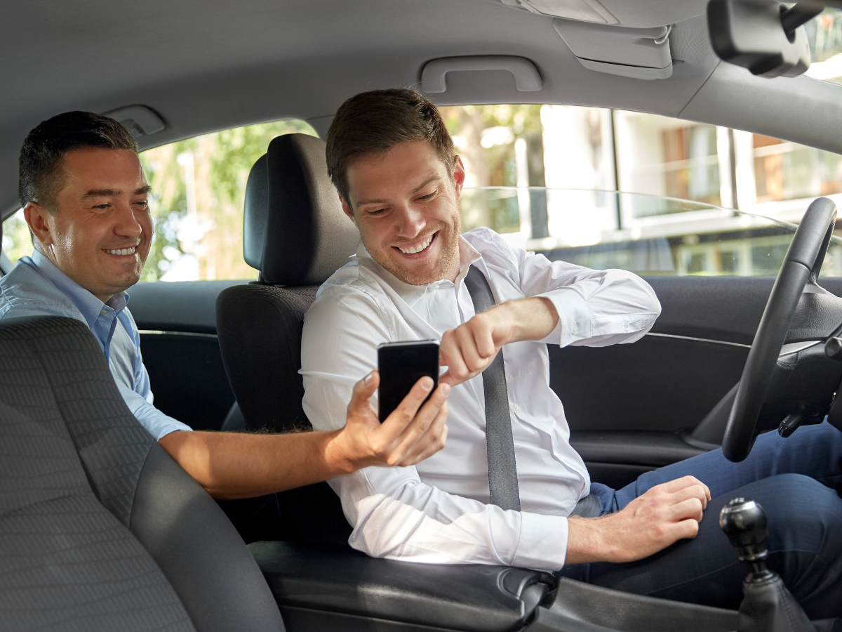 Two men in a car taking a selfie, one smiling, the other holding the phone. Interior shot.