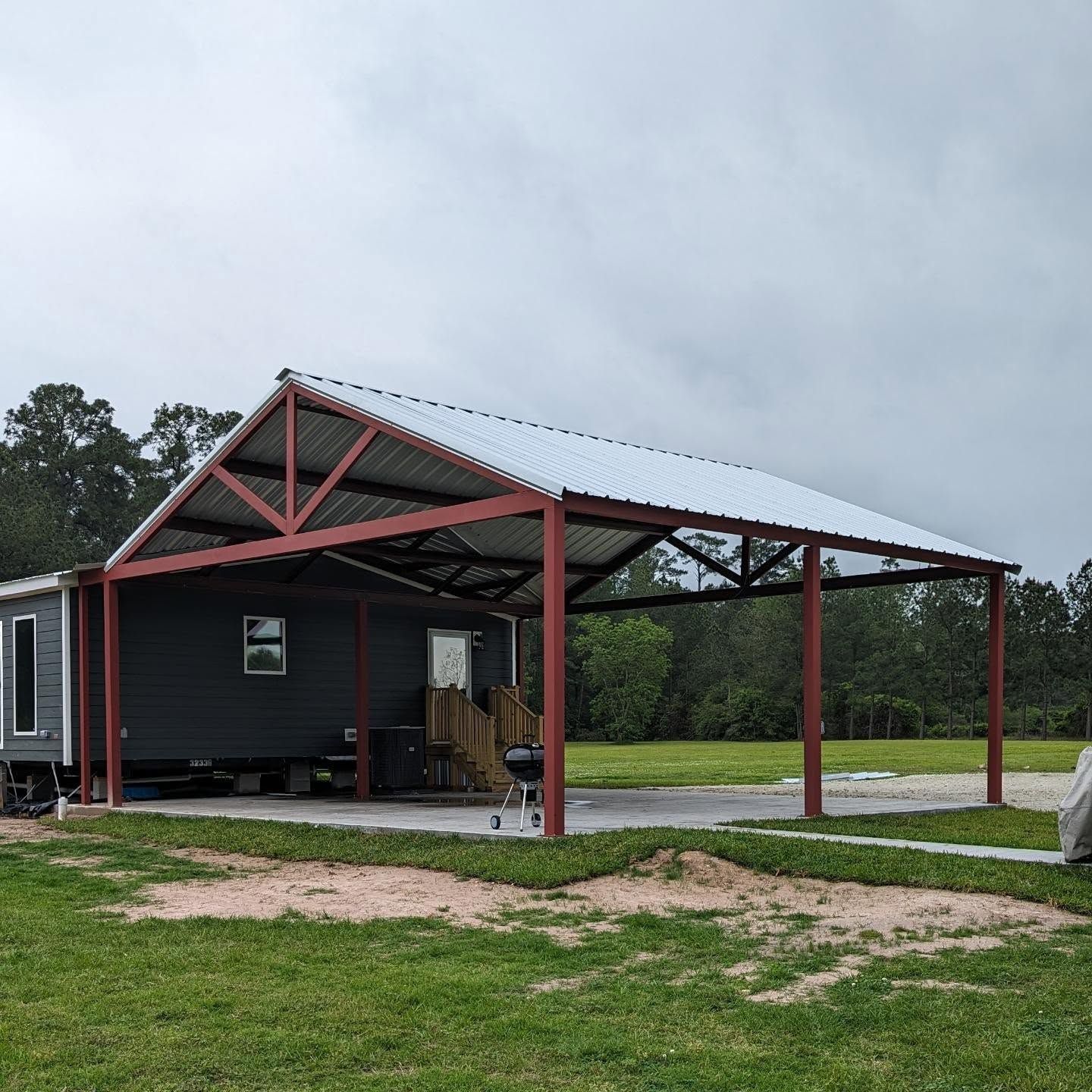 Red metal carport attached to a gray mobile home. Metal roof, concrete pad, green grass, and overcast sky.
