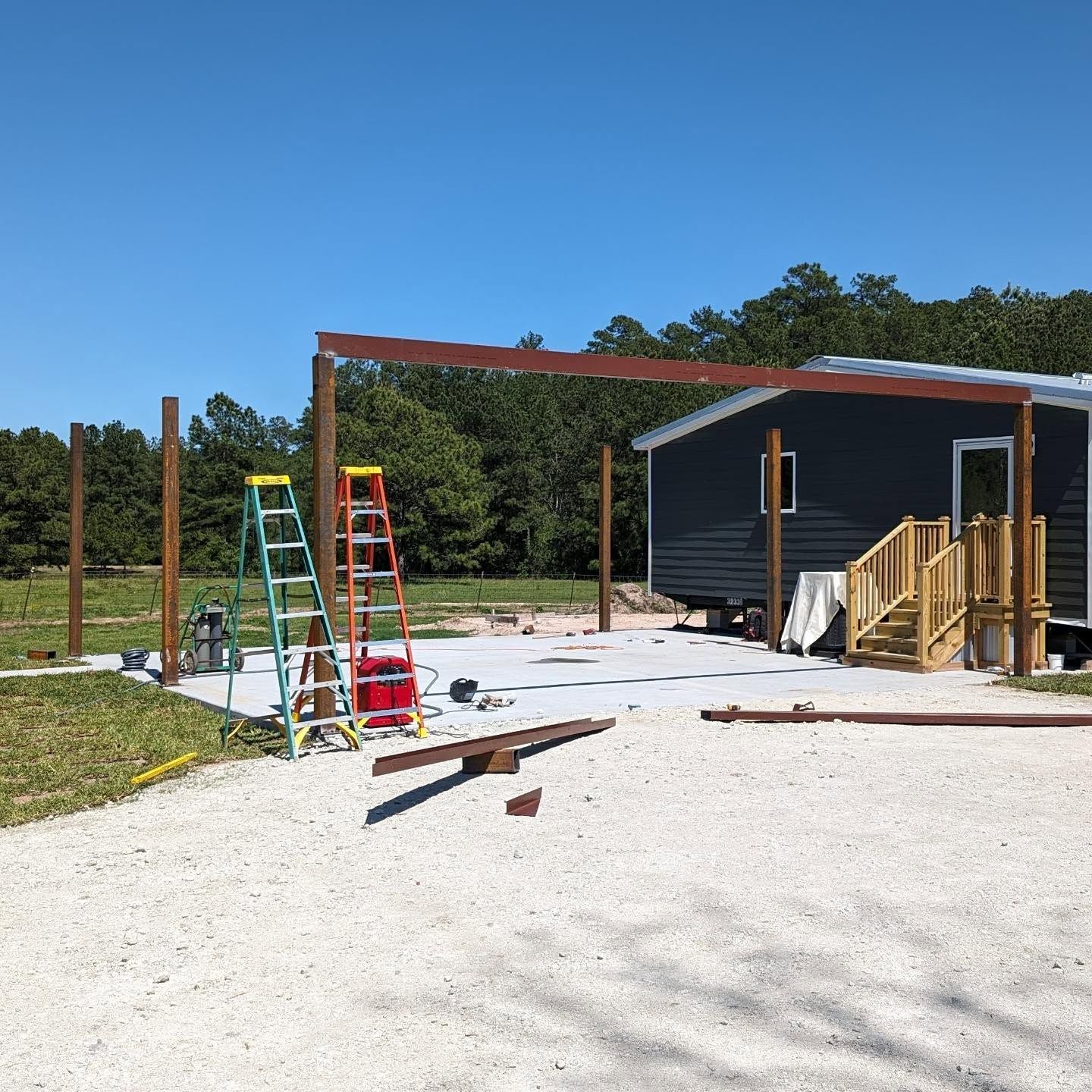 Construction of an open-air carport attached to a dark blue house on a concrete pad with two ladders.