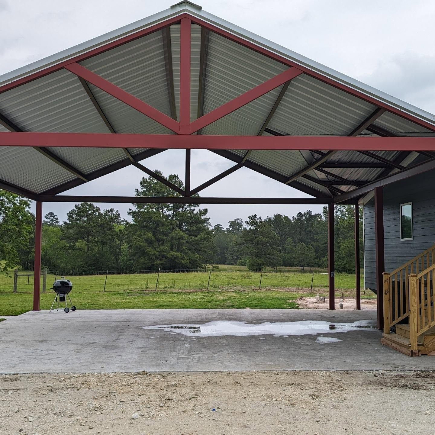 Red metal carport with gray roof, concrete pad, grill, and view of trees.