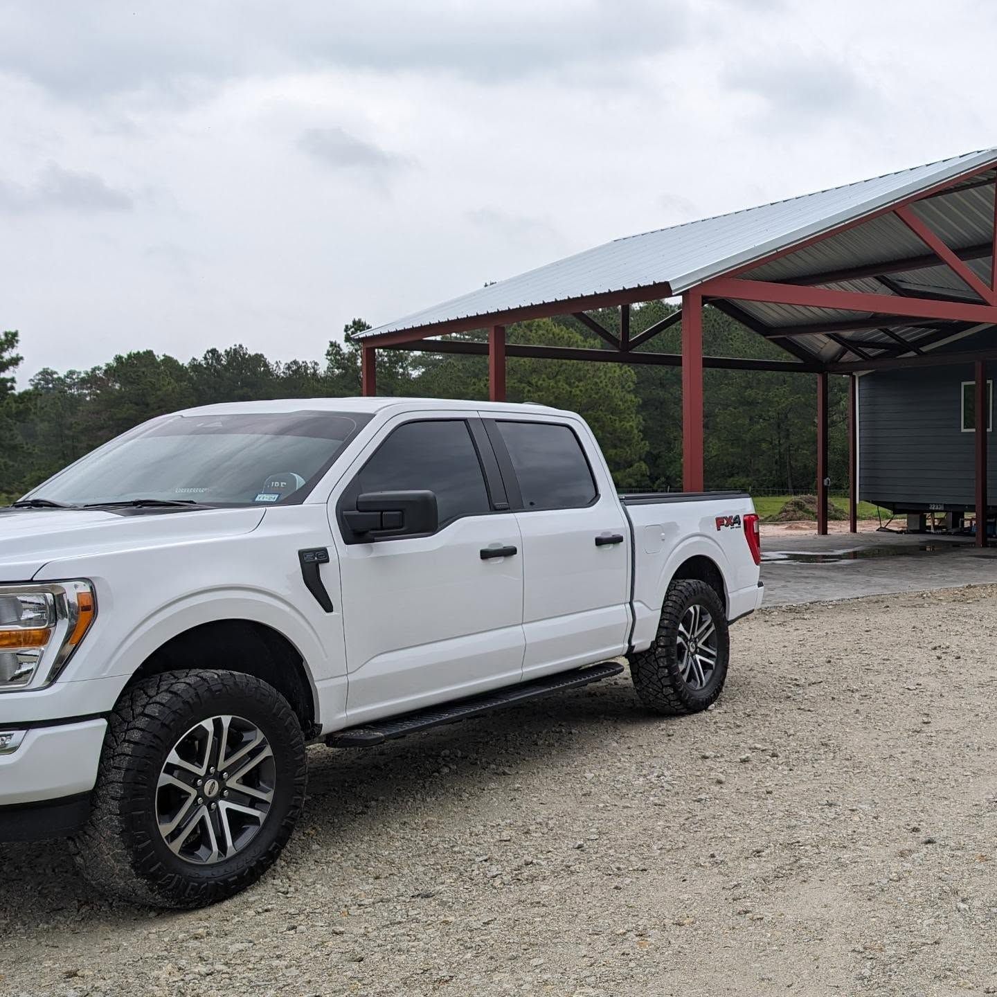 White pickup truck parked under a metal roof structure. Gravel driveway, trees in background.