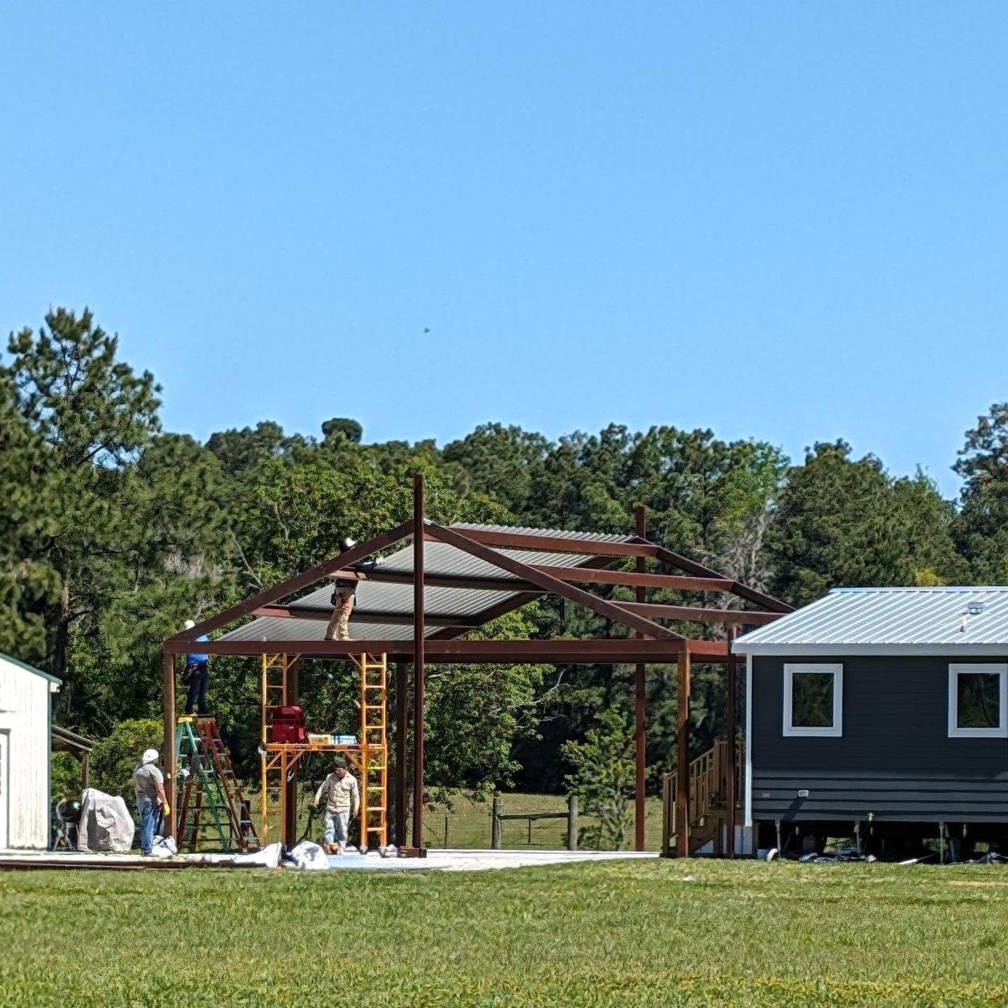 Construction of a brown-framed roof structure, workers on scaffolding, blue sky, green grass.
