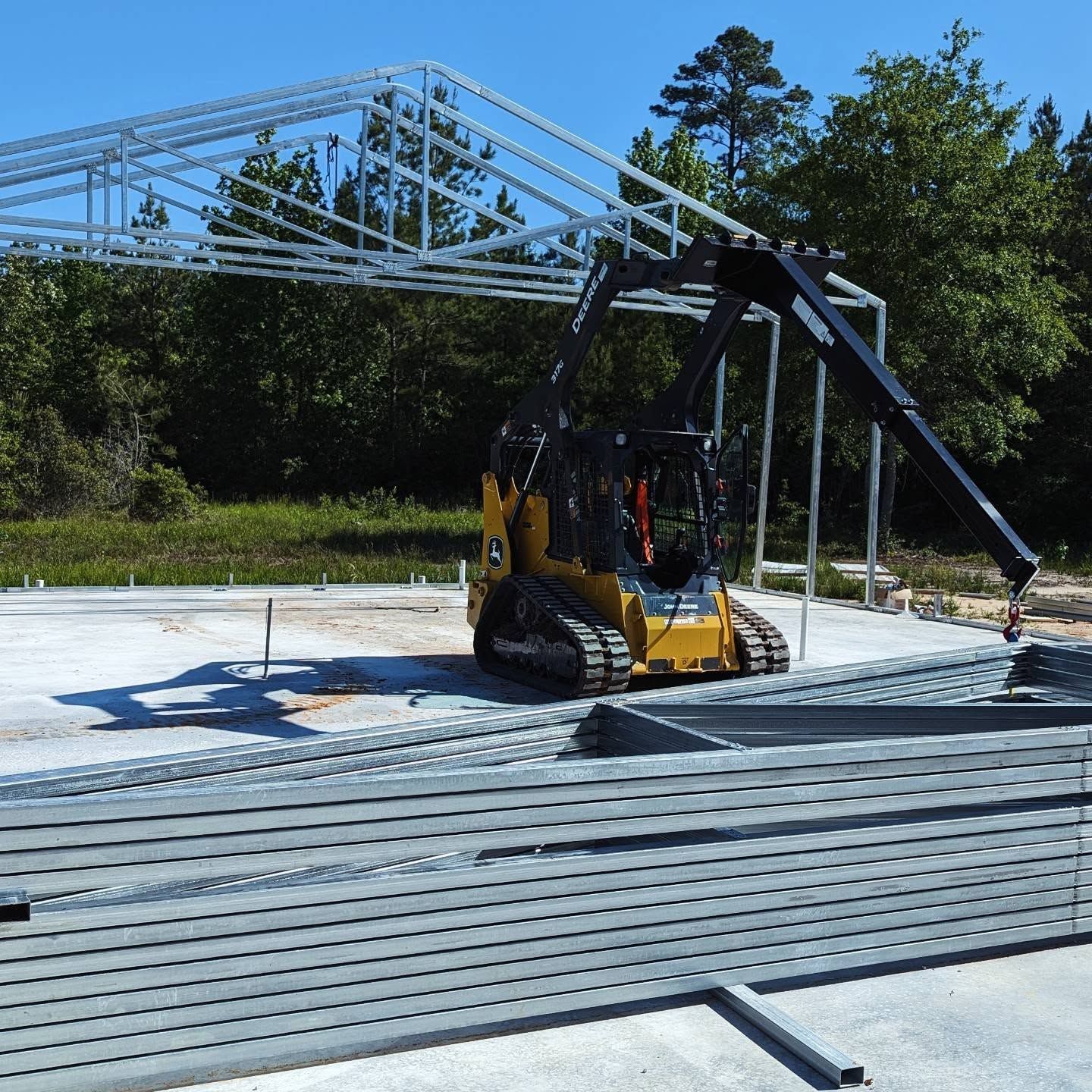 Yellow skid steer with crane arm lifting metal beams at a construction site.