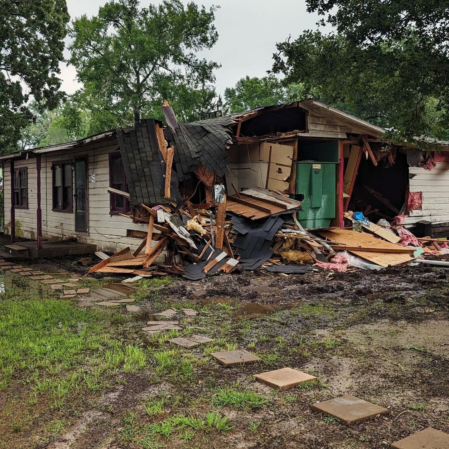 House severely damaged with collapsed roof and debris in the yard. White siding, green door, overcast sky.