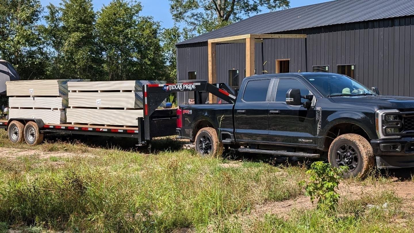 Black truck towing a trailer loaded with lumber near a dark building and greenery.