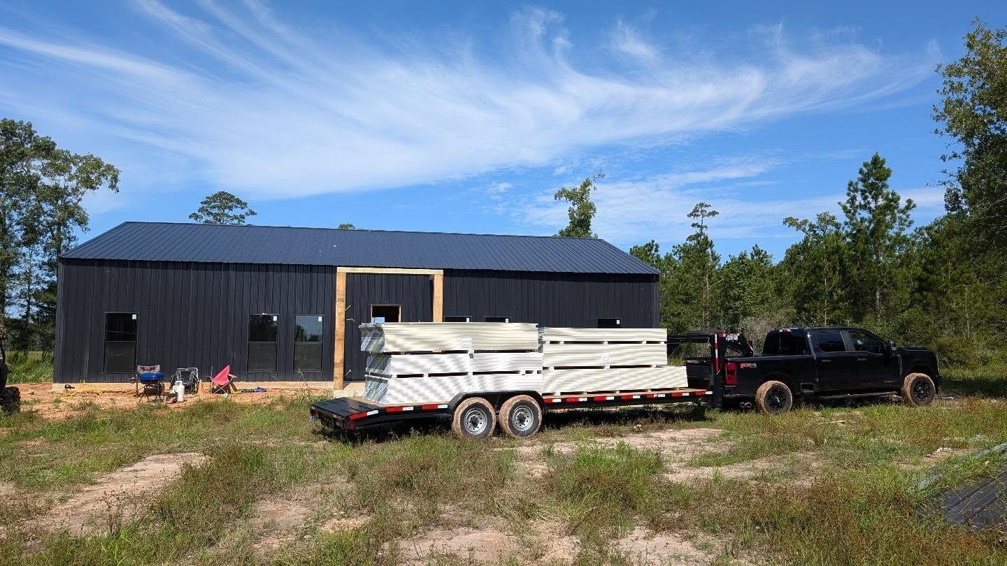 A black house under construction with a truck towing a trailer of building materials on a sunny day.