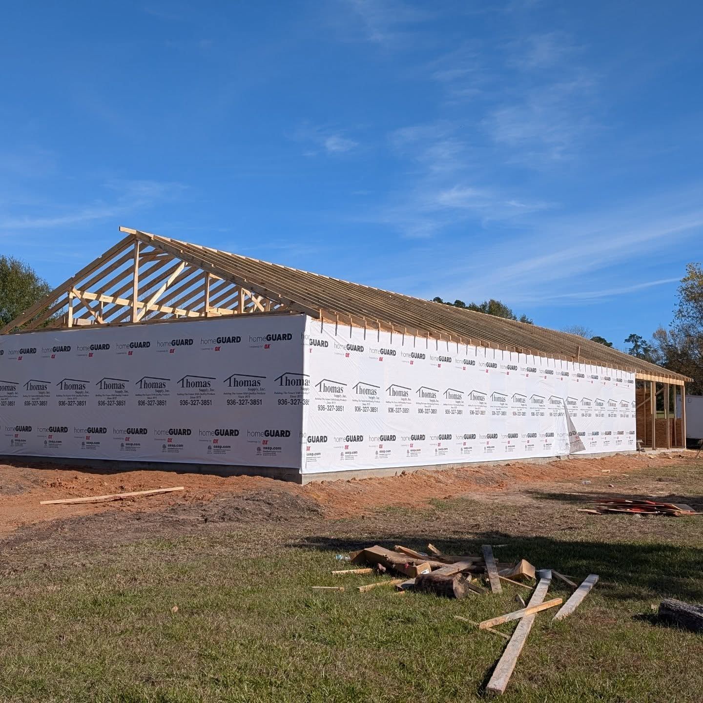 Building under construction: wood frame with roof trusses, sheathing, and wrapped walls against a blue sky.