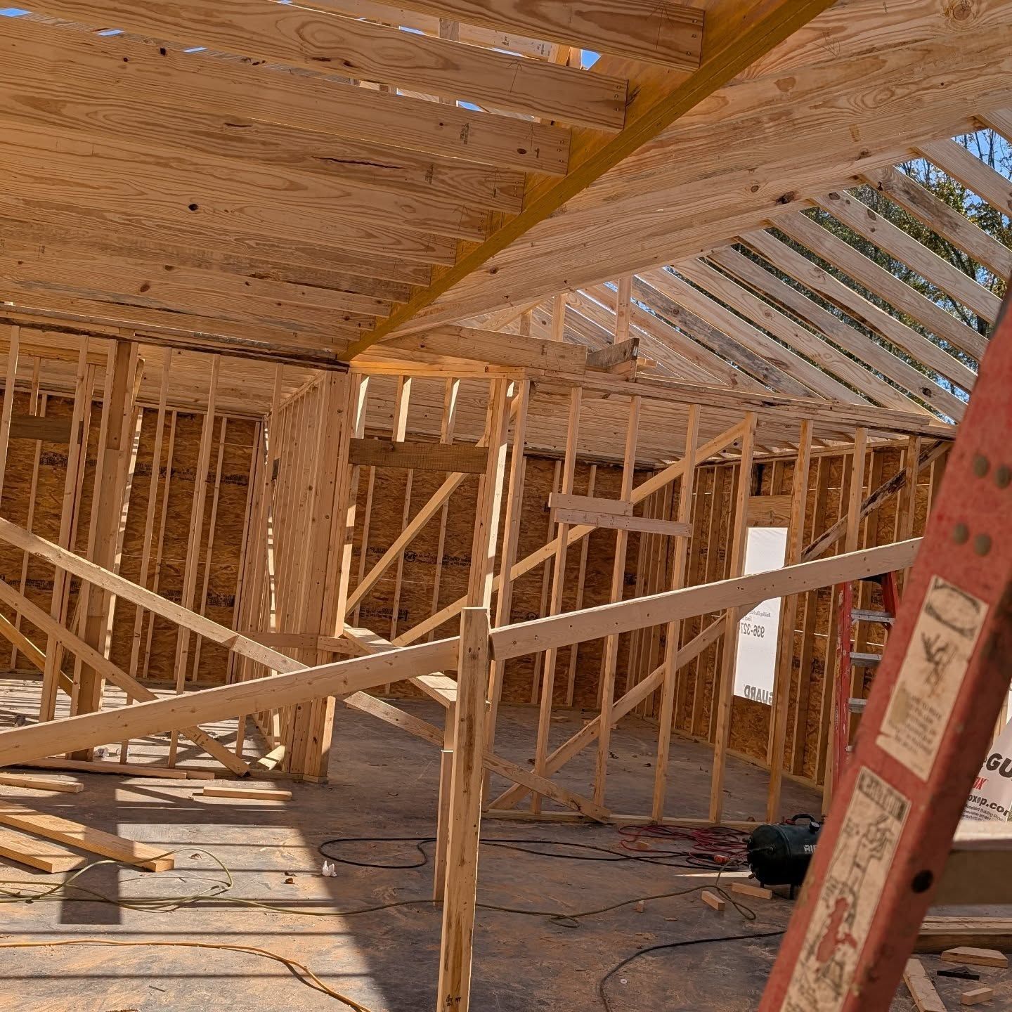 Wood framing of a building under construction. Studs, rafters, and a ladder are visible. Sunlight streams in.