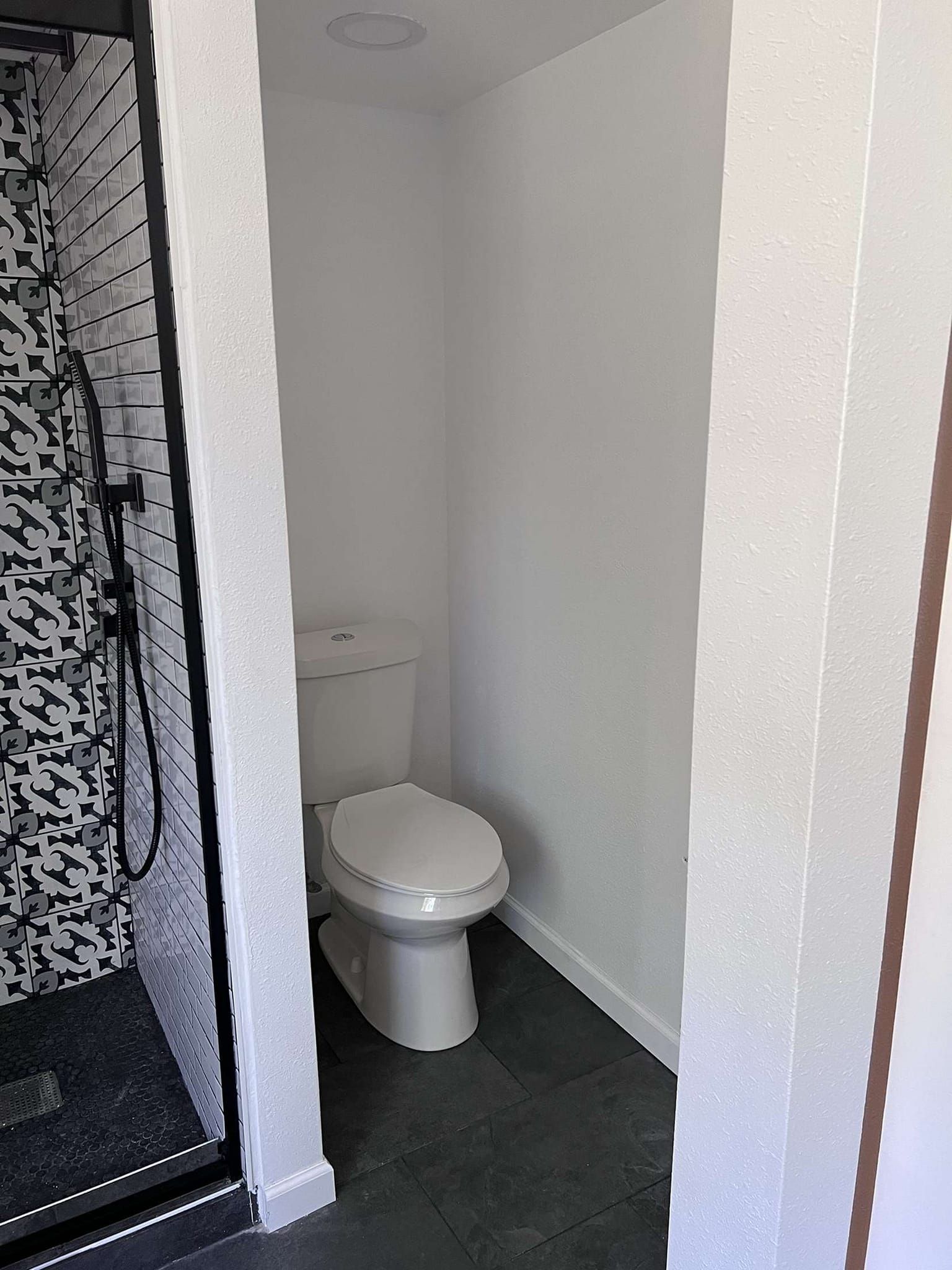 Small bathroom with a toilet and shower visible. White walls, black floor, and mosaic tile in shower.