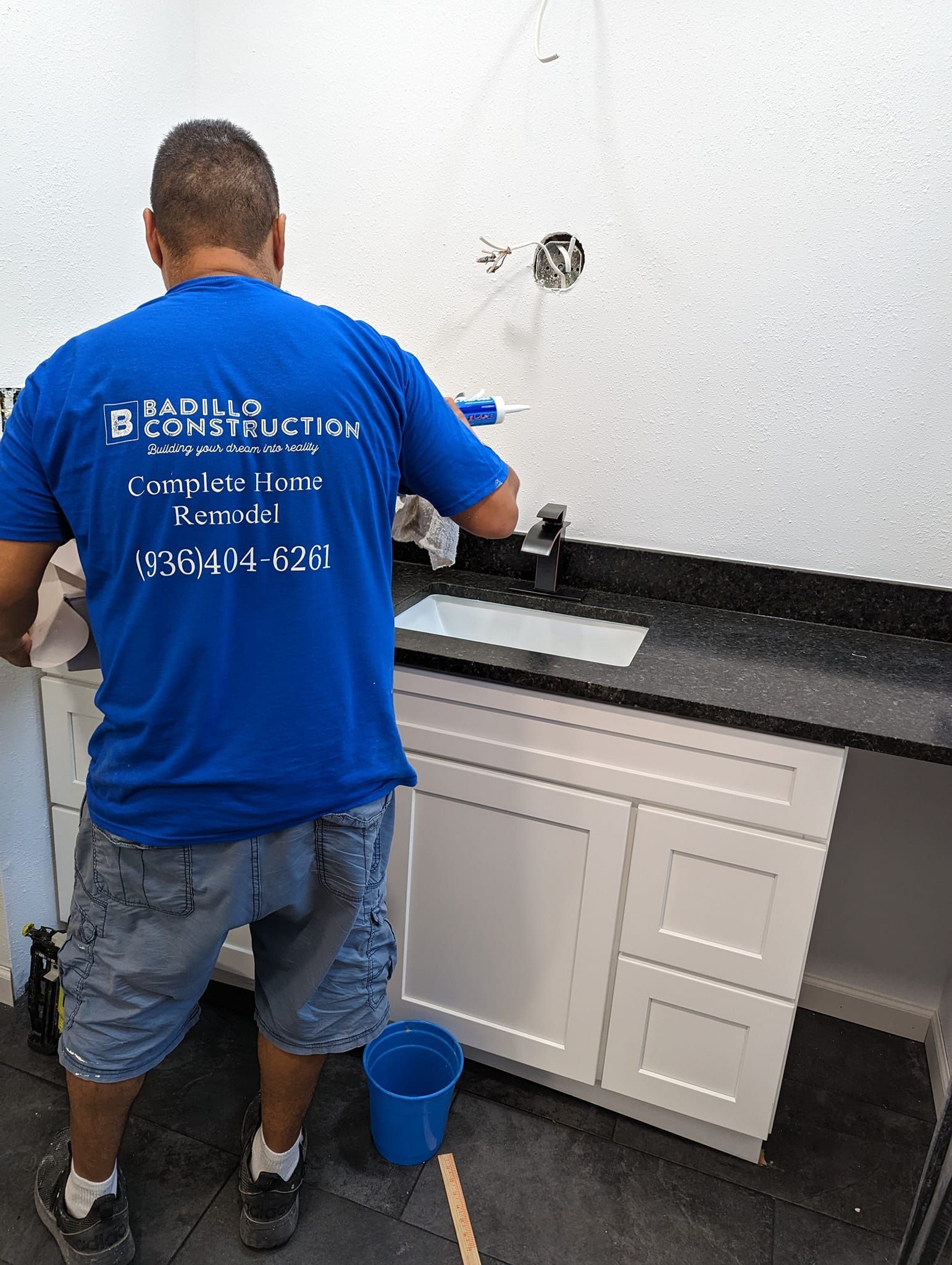 Man in blue shirt installing a bathroom vanity; white cabinets, black countertop, blue bucket.