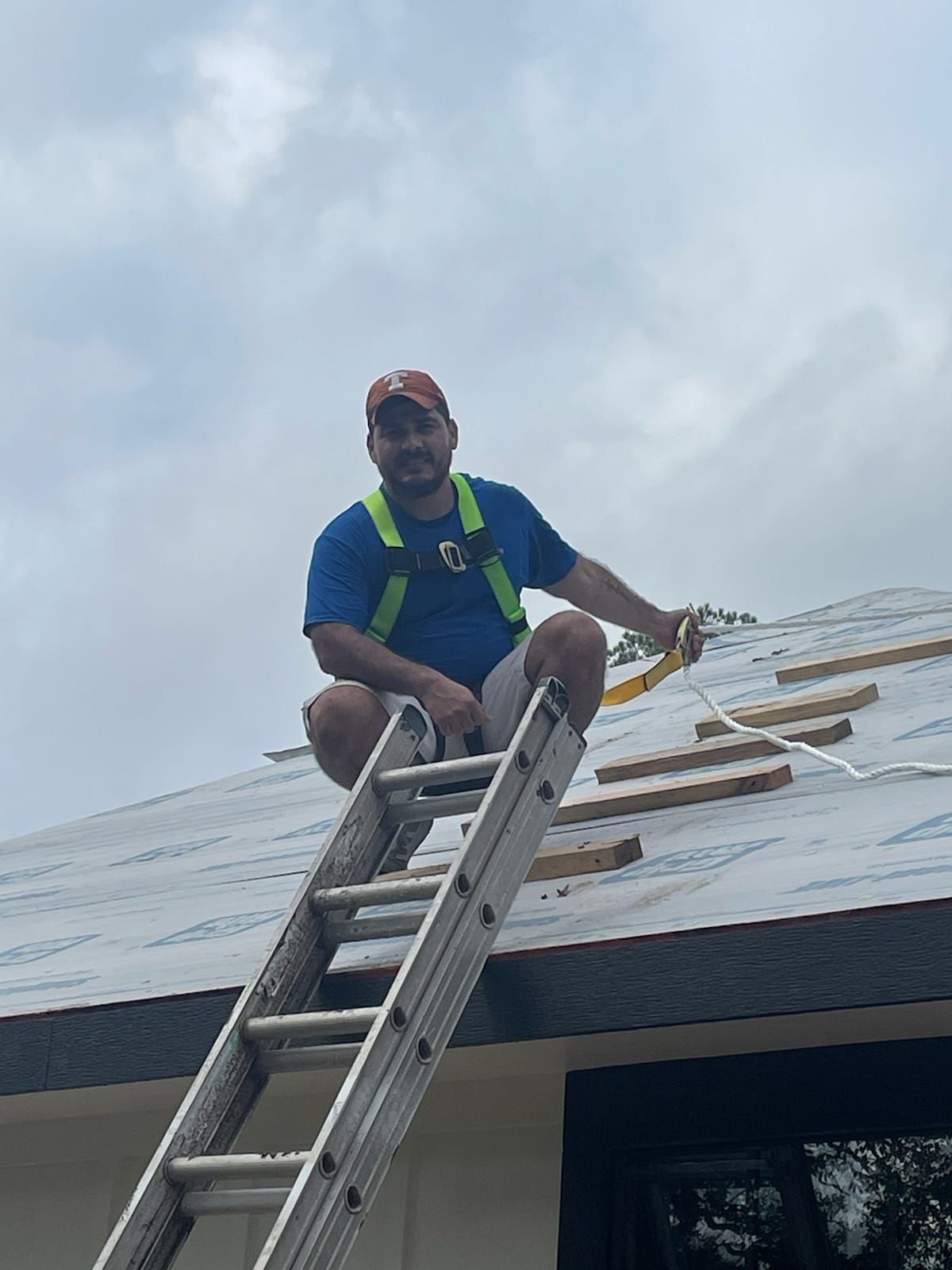 Man on roof, wearing safety harness, seated on ladder, installing roofing materials. Overcast sky.