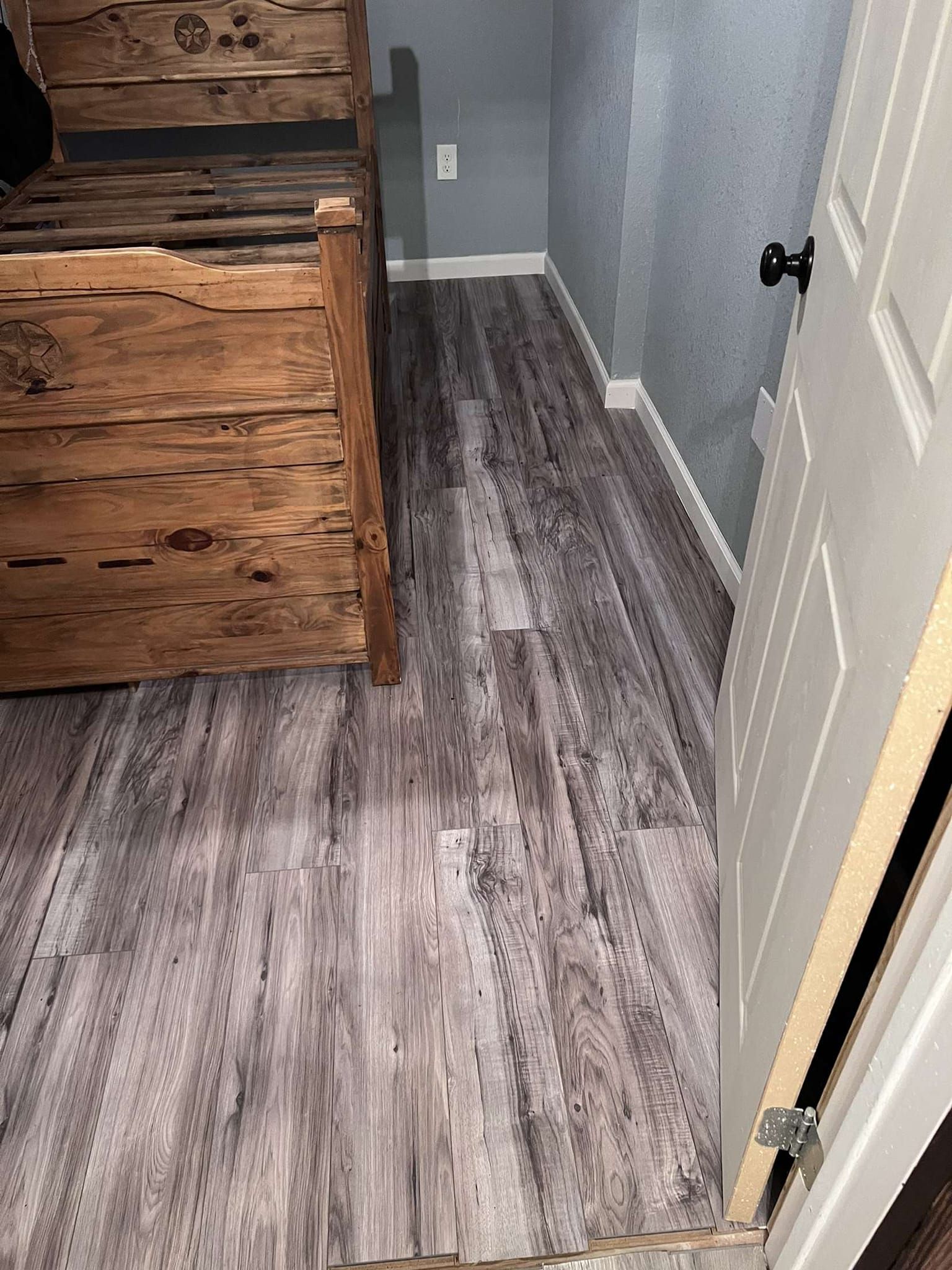 Wood-look flooring in a room with a rustic bed frame, grey walls, and a white door.