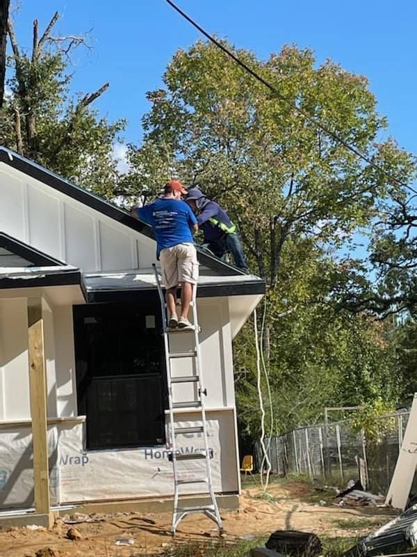 Two workers on a roof, one on a ladder, installing something on the edge of a newly built house.