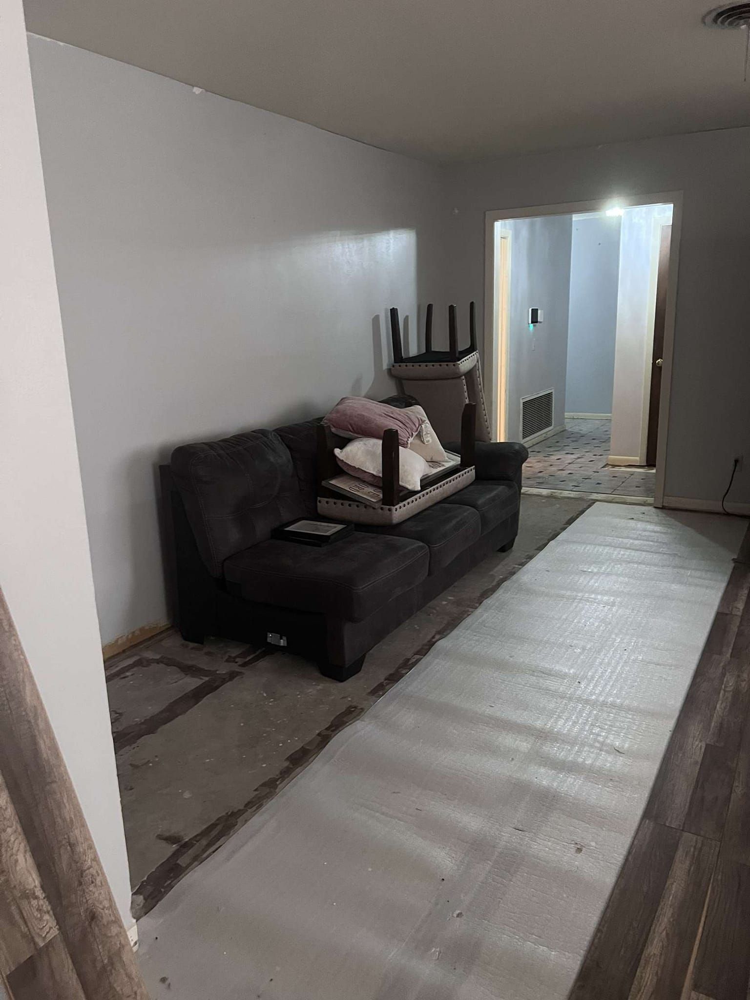 Gray sofa in a room with white walls, open doorway, and flooring under renovation.