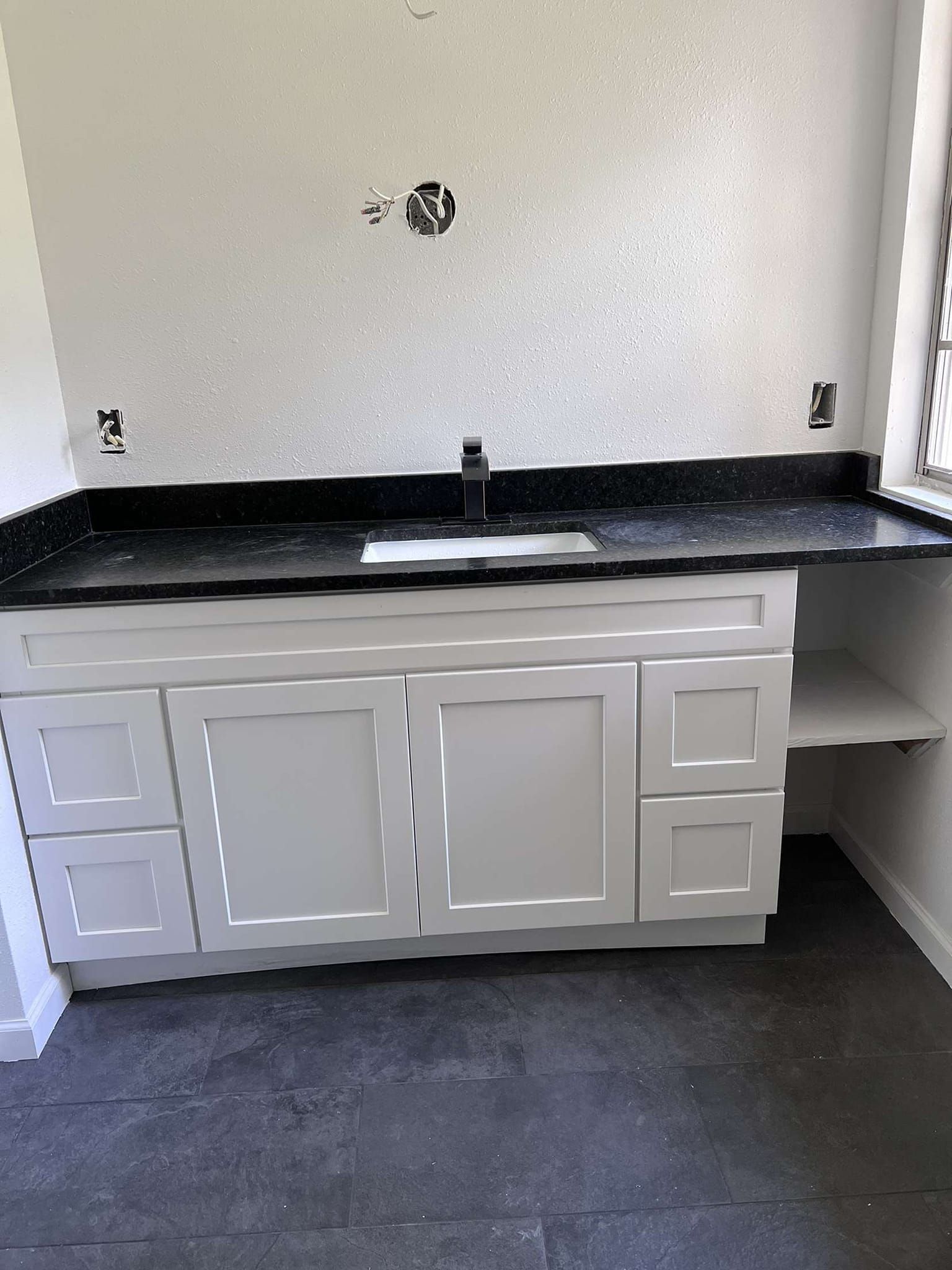 White bathroom vanity with black countertop, faucet, and sink against a textured white wall.