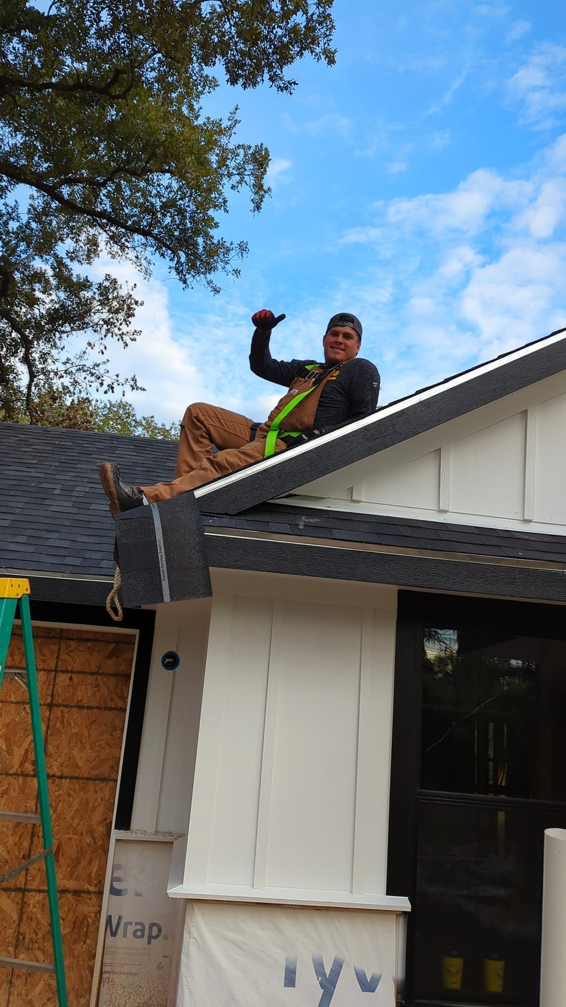 Person on rooftop gives thumbs-up, wearing a safety harness. Black shingle roof, white siding, blue sky.