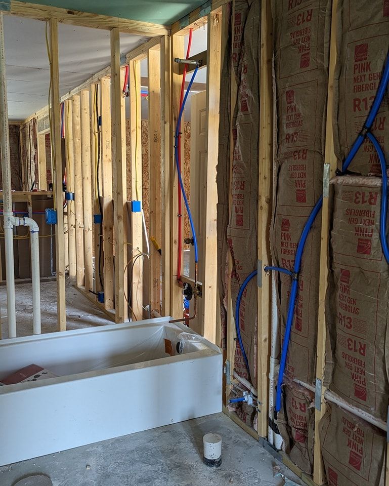 Bathroom under construction, showing wooden studs, blue water pipes, insulation, and a white bathtub.