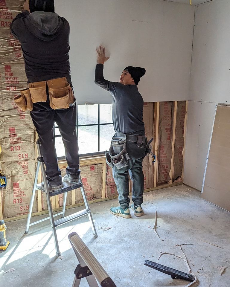 Two people installing drywall in a room with exposed wall studs and insulation.