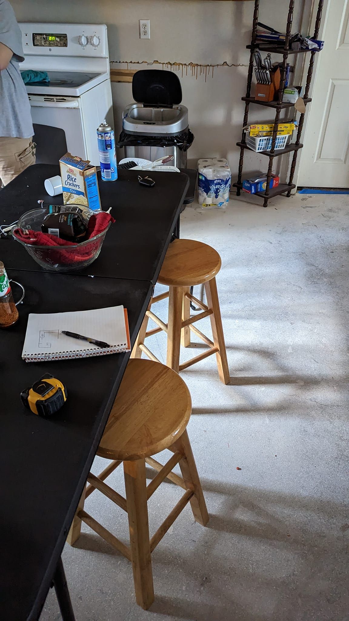 A kitchen with a long black table, two wooden stools, a stove, and items on shelves.