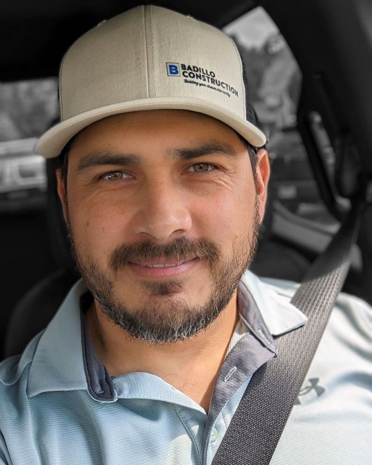 Man wearing a hat with the Bazelio Construction logo smiles, inside a vehicle, wearing a seatbelt.
