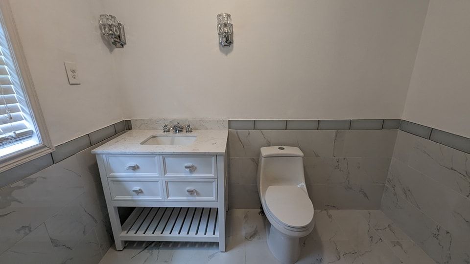 White bathroom with marble tiles, vanity, and toilet. Two sconces above the toilet and vanity.