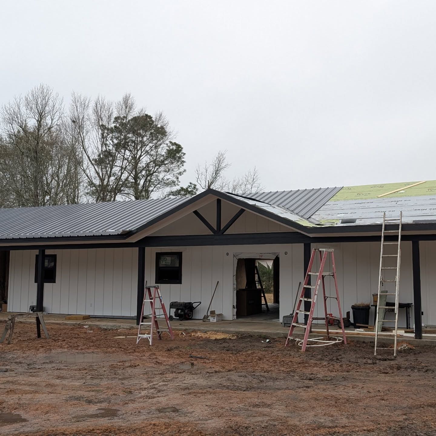 A one-story building under construction with white siding, black trim, and ladders. The roof is partially covered with dark tiles.