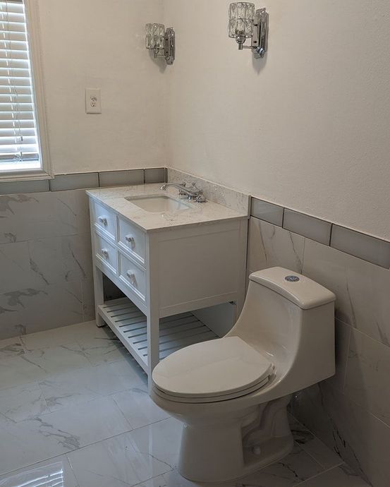 White bathroom with vanity, toilet, and marble-look tile. Two sconces hang above the vanity.