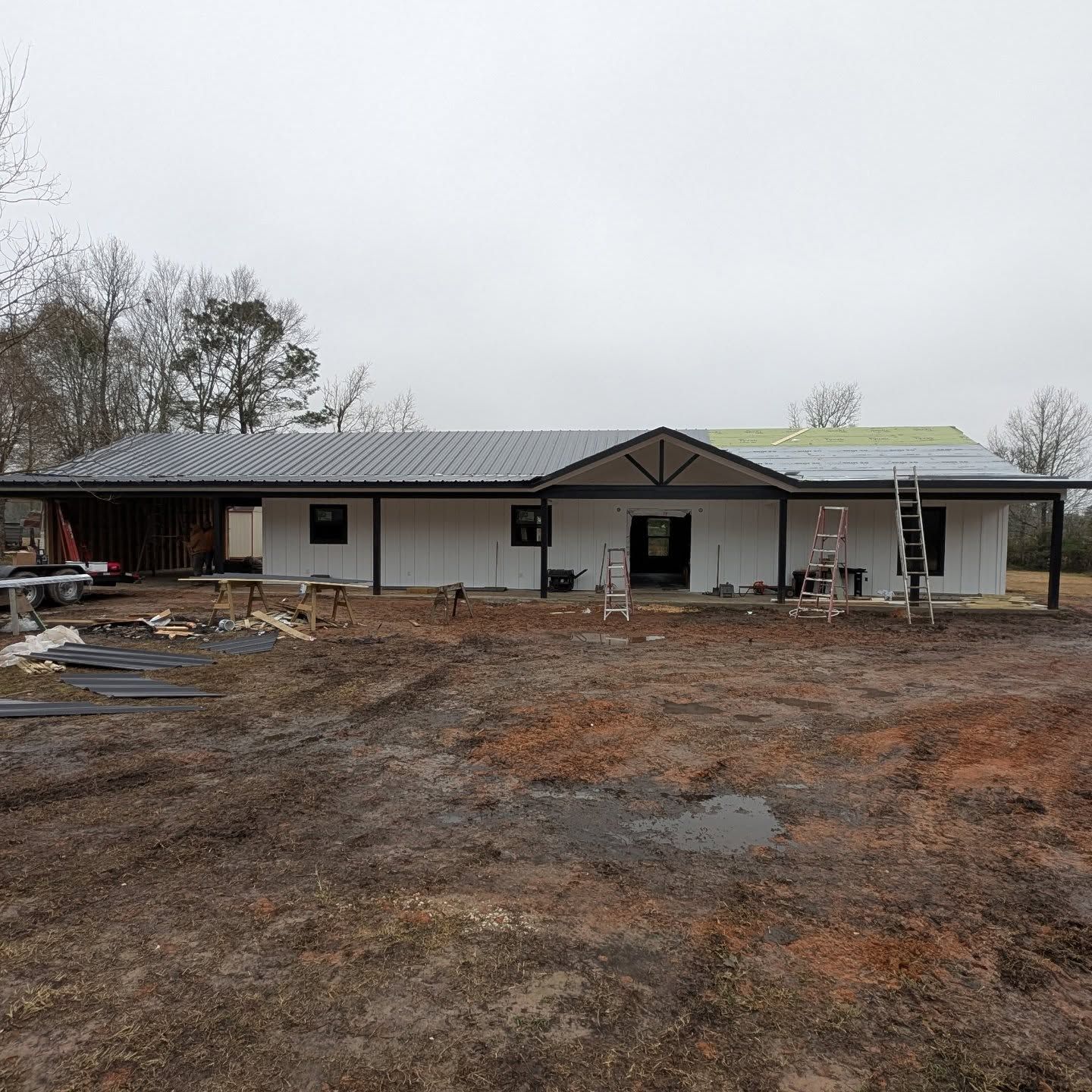 A one-story building under construction with white siding, a black metal roof, and a covered porch.