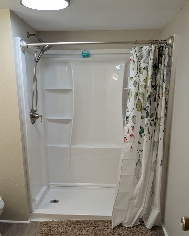 White shower stall with floral shower curtain, silver showerhead, and a brown rug.