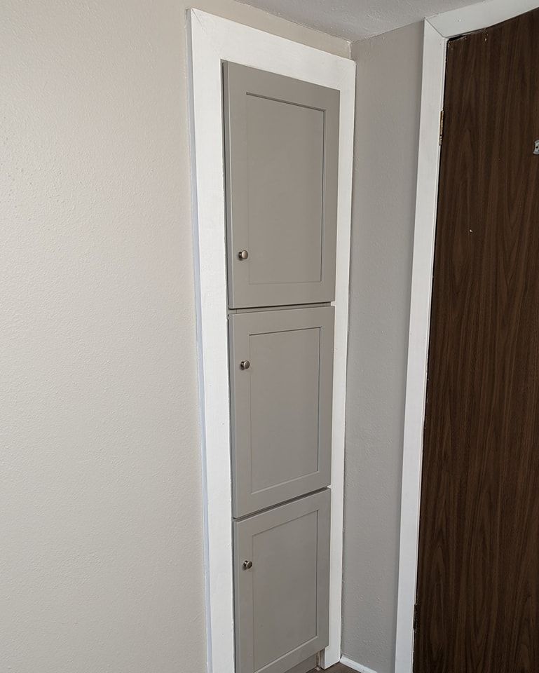 Three-door gray storage cabinet recessed in a wall. White trim surrounds it and a brown door is beside it.
