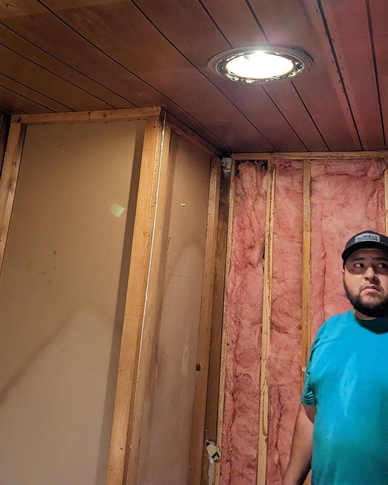 Man in blue shirt stands near open wall framing with exposed pink insulation. Wooden ceiling with recessed light.