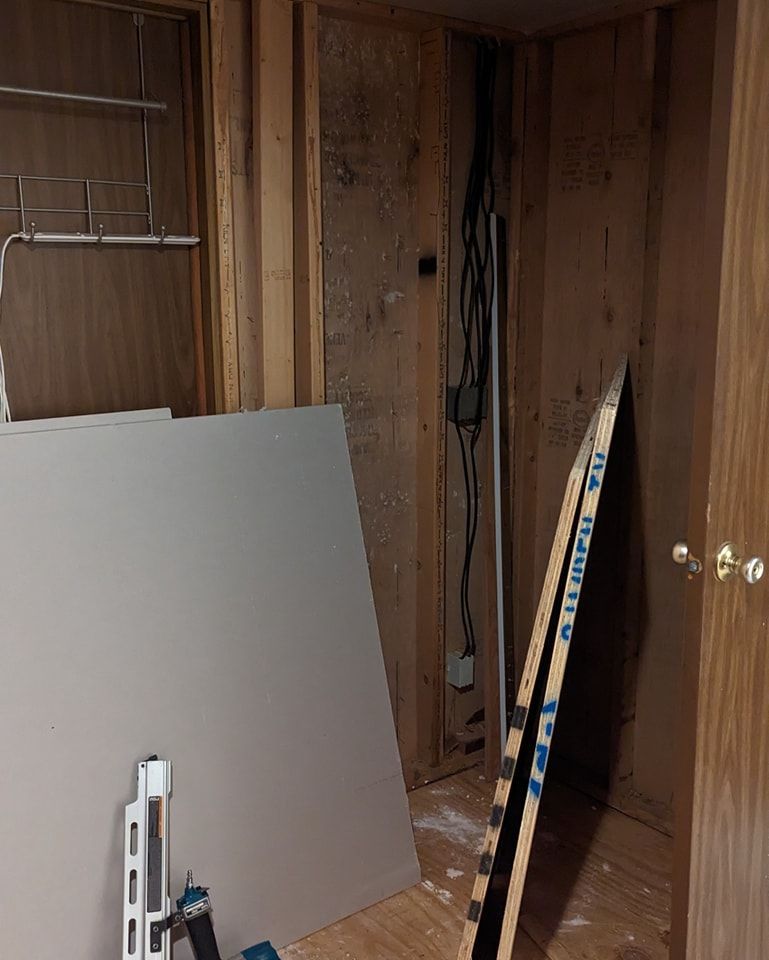 Interior wall under construction; drywall and boards lean against the wood frame. Wires hang in the corner.
