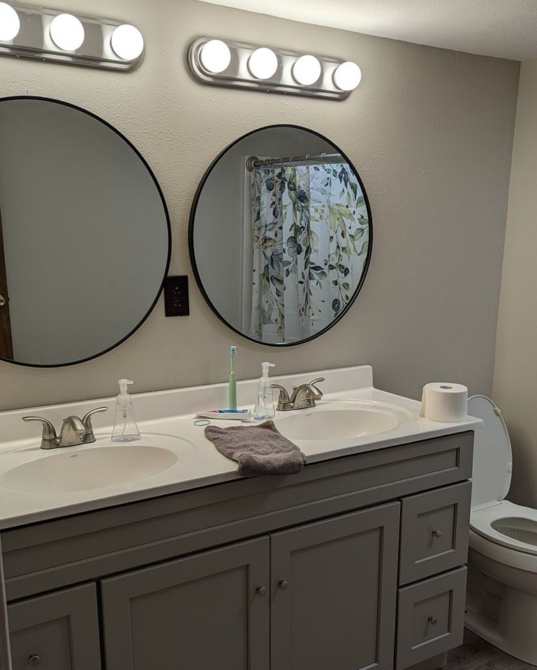 Bathroom with double vanity, two round mirrors, gray cabinets, and a toilet.