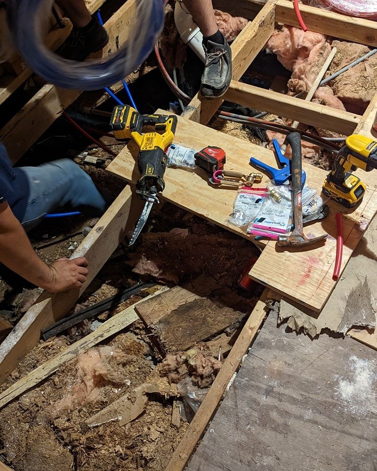 Construction workers in crawlspace, using tools and materials for plumbing, including saws and pipes.