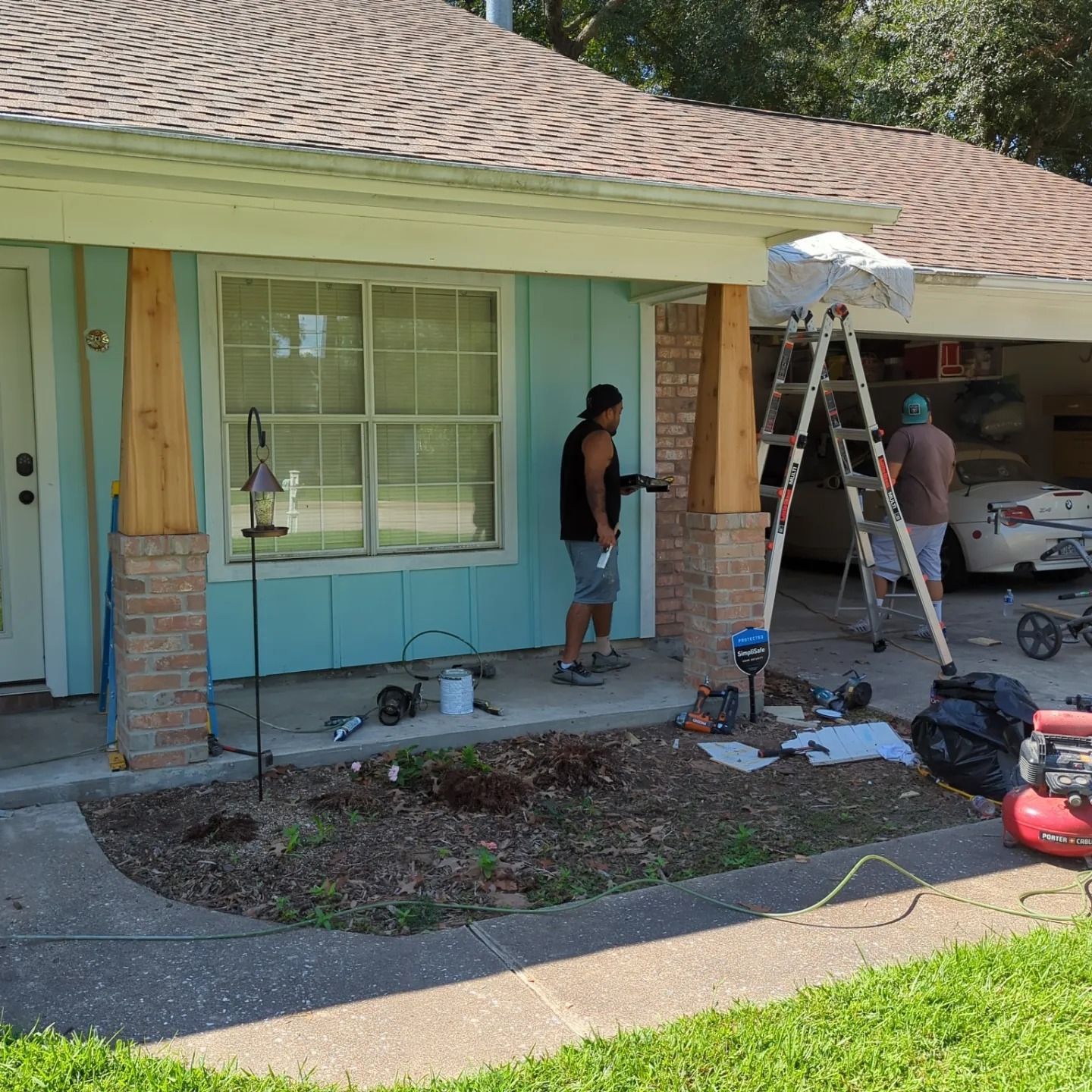 Two people working on a house exterior. One uses a nail gun on a wooden post. Another stands on a ladder.