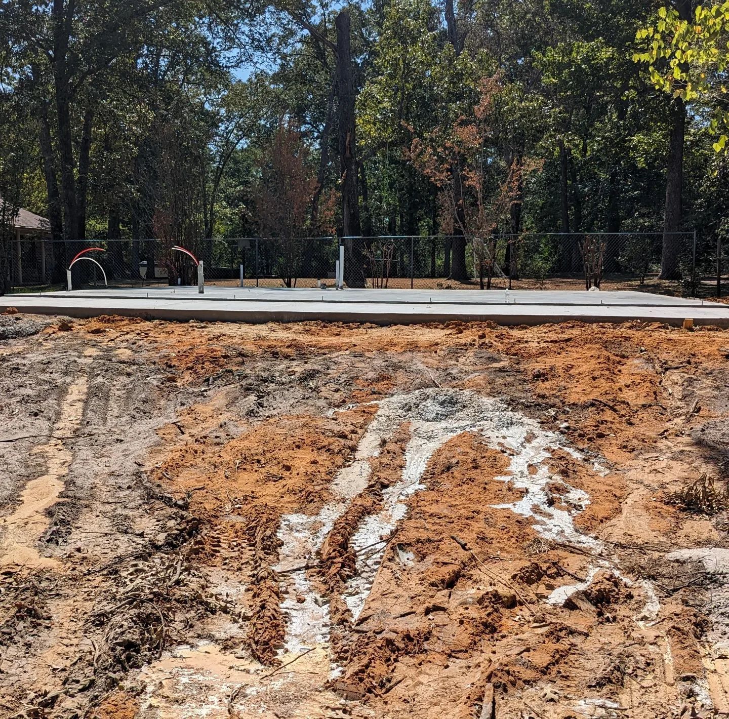 Dirt and mud in foreground, paved area with playground equipment in background, trees, sunny day.