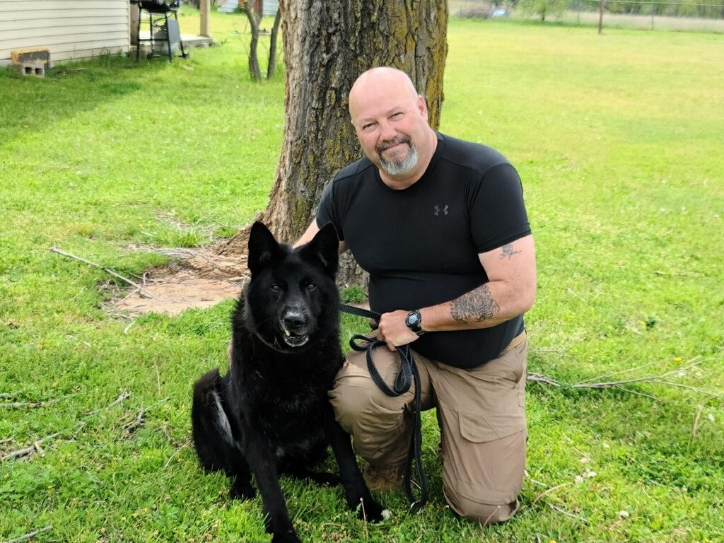 A man is kneeling down next to a black dog on a leash.