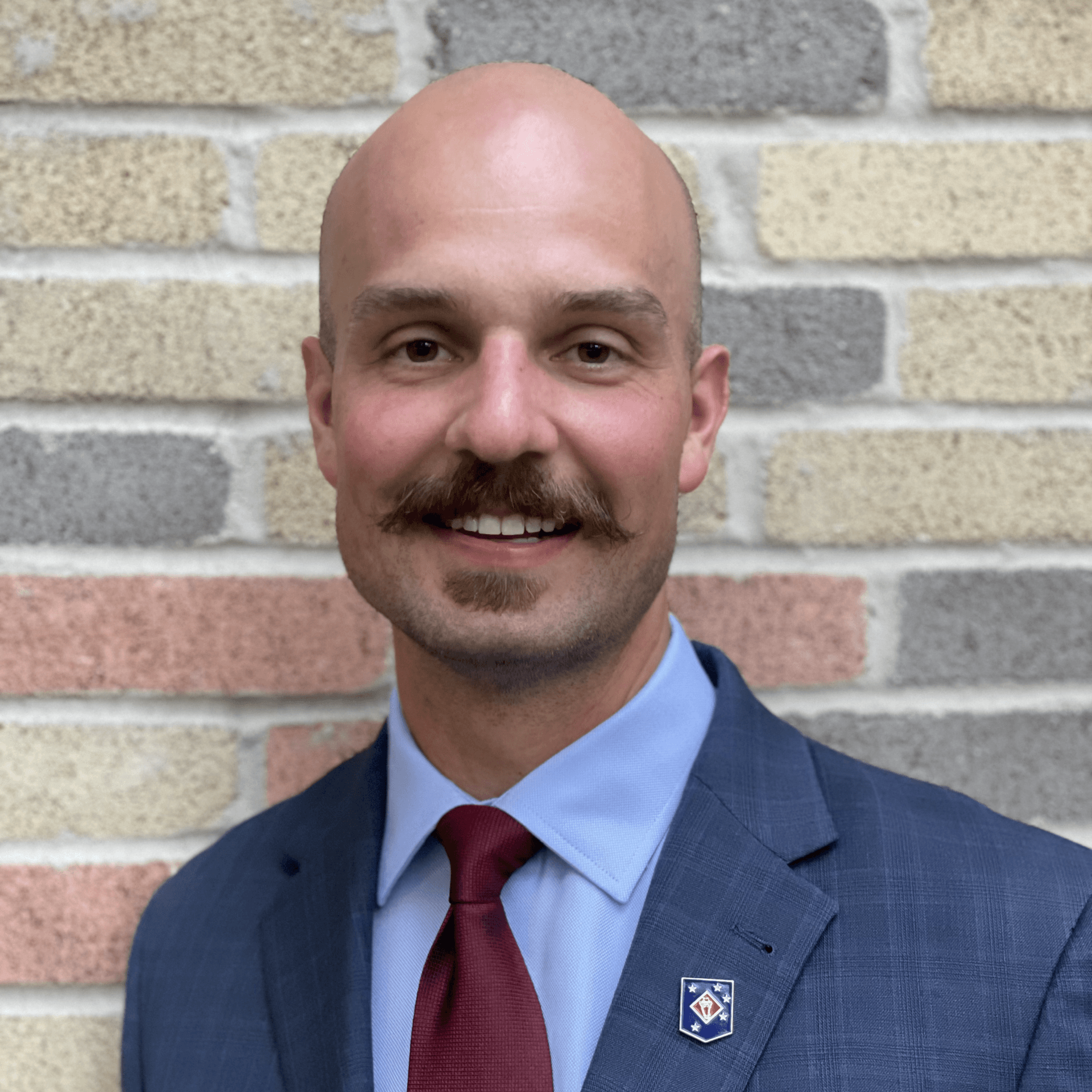 A bald man with a mustache wearing a suit and tie is smiling in front of a brick wall.