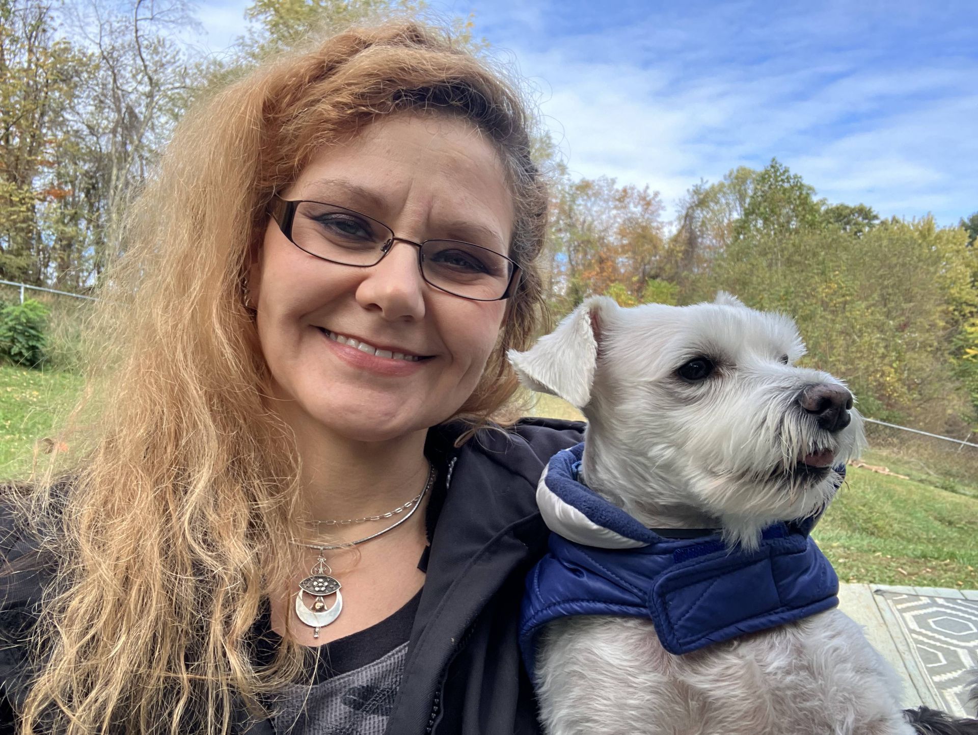 A person with wavy blonde hair and glasses smiles while holding a small, white dog wearing a blue vest outdoors.