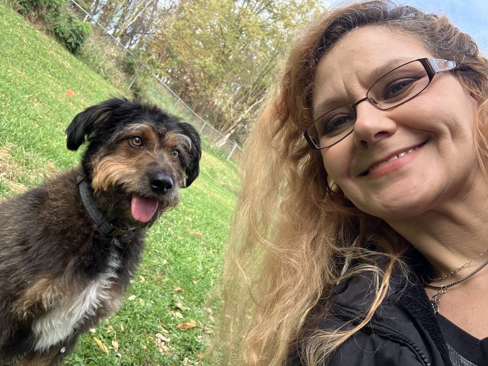 A woman with curly reddish-blonde hair and glasses smiling in a park next to a happy, brown and black scruffy dog.