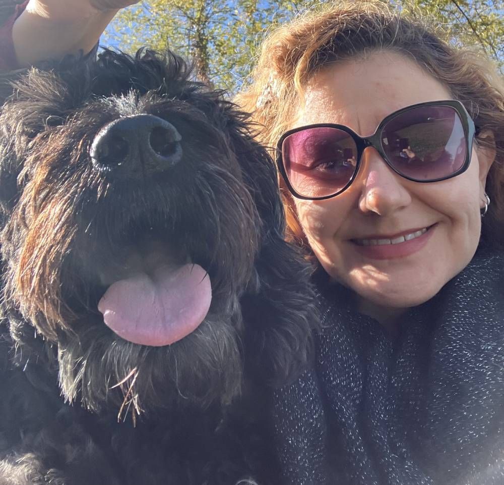 A woman wearing sunglasses smiling next to a happy, long-haired black dog with its tongue out, outdoors in the sun.