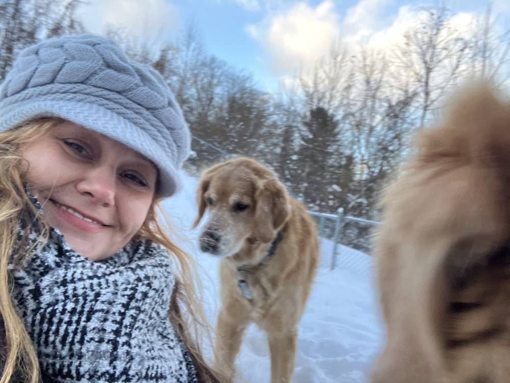 A smiling person wearing a grey knit hat and a black-and-white scarf poses with two dogs in a snowy outdoor setting.