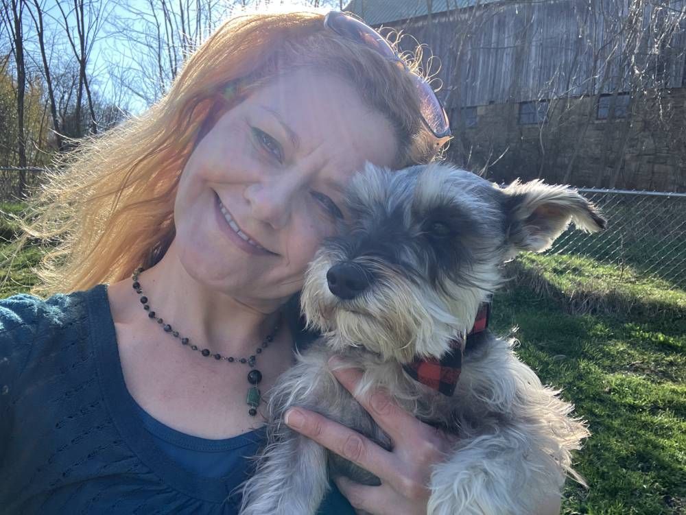 A smiling person holds a scruffy, grey miniature schnauzer wearing a red and black plaid bowtie outdoors in the sun.