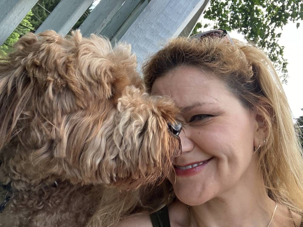 A person smiling closely next to a curly, light-brown dog outdoors.